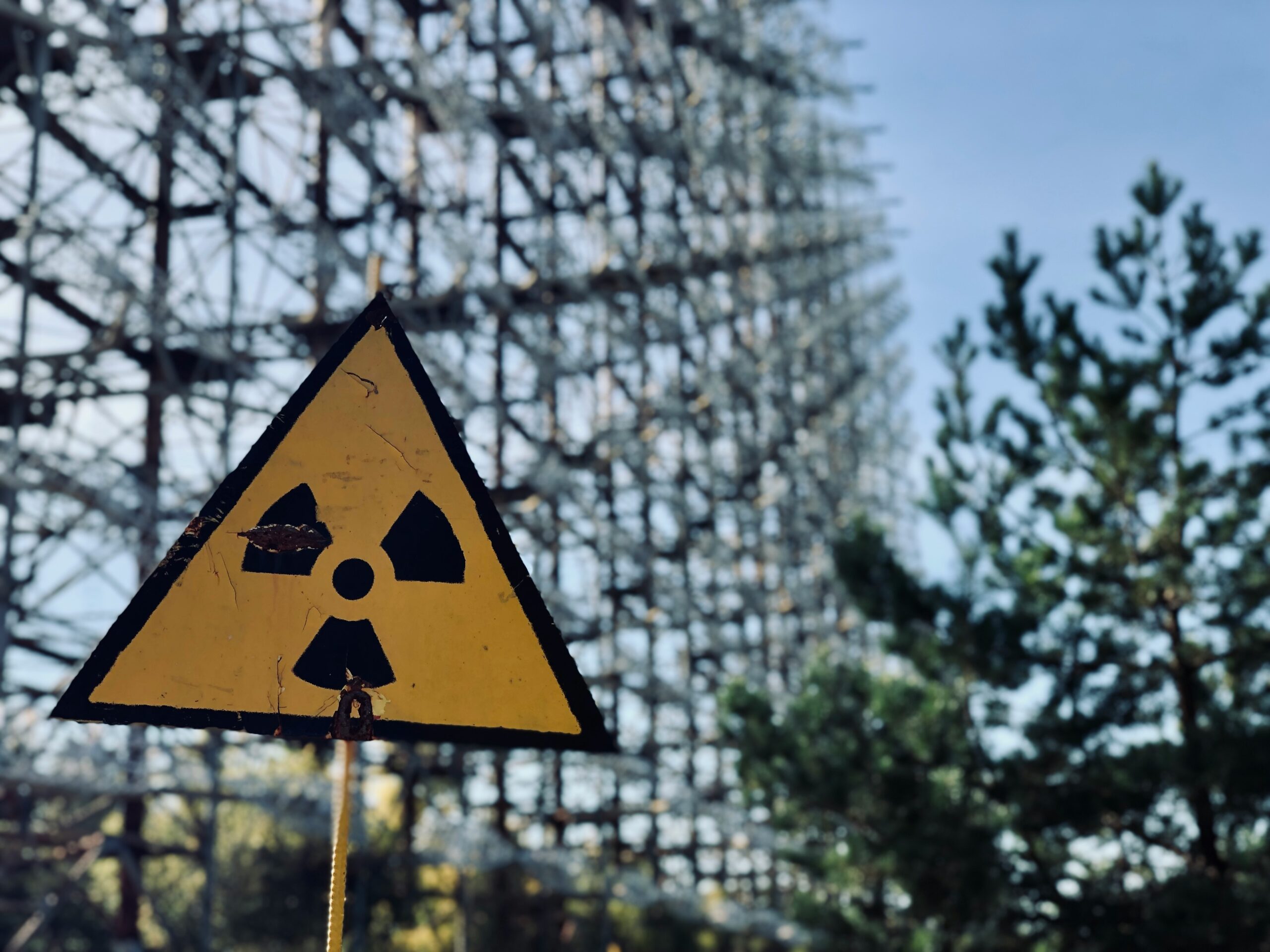 A yellow radiation warning sign in the Chernobyl Exclusion Zone, with the abandoned Duga-3 radar complex visible in the background.
